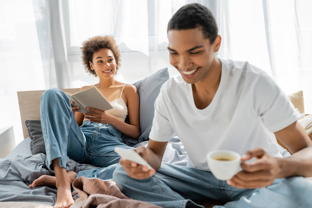 Neşeli Afro-Amerikalı kadın elinde kitapla yatakta oturmuş ön planda kahve ve cep telefonu olan sevgilisine bakıyor. - Fotoğraf, Görsel