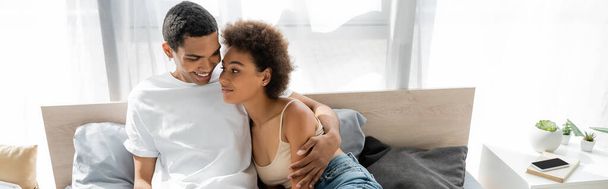 joyful african american man in white t-shirt hugging curly girlfriend on bed, banner - Photo, Image
