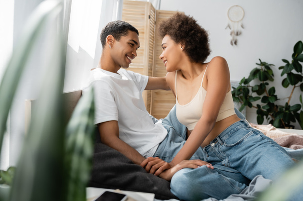 sexy african american couple sitting on bed and smiling at each other on blurred foreground - Foto, immagini
