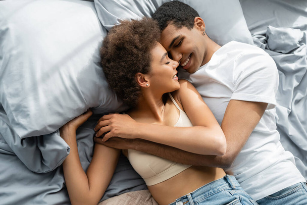 top view of african american woman in crop top and man in t-shirt hugging with closed eyes on bed - Foto, immagini