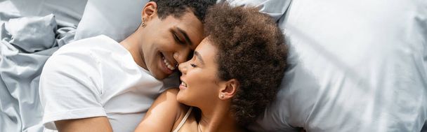 pohled shora na mladý africký americký pár ležící na posteli a usmívající se tváří v tvář se zavřenýma očima, prapor - Fotografie, Obrázek