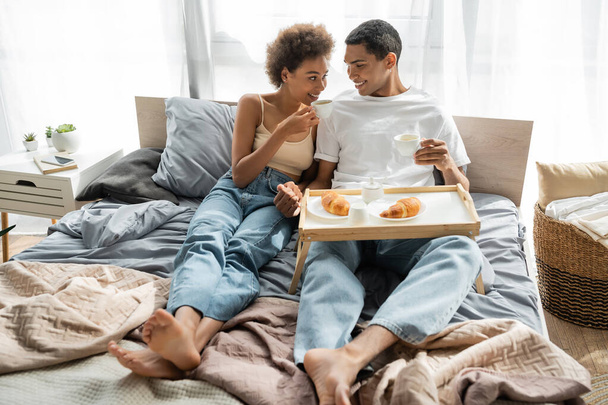 молодая африканская американская пара в джинсах пьет кофе возле подноса с круассанами на кровати - Фото, изображение