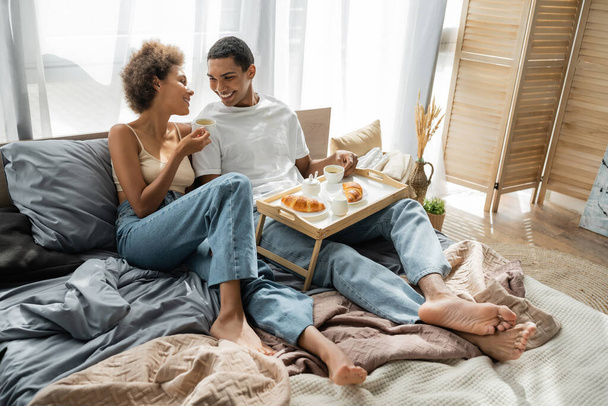 full length of barefoot african american couple in jeans sitting near tray with coffee and croissants on bed - Foto, afbeelding