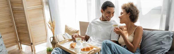 young and happy african american couple smiling near breakfast with coffee and croissants on bed, banner - Photo, Image