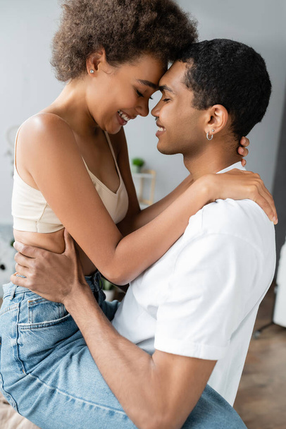 side view of african american man in white t-shirt and sexy woman in crop top hugging with closed eyes at home - Foto, afbeelding