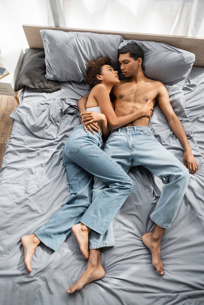 wysoki kąt widzenia boso afrykańskiej pary amerykańskiej w dżinsach leżących na łóżku i patrzących na siebie  - Zdjęcie, obraz
