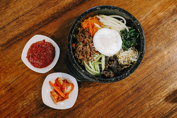 Flache Lage- oder Draufsicht auf die koreanische Küche, die als Bibimbap oder Dolsot bekannt ist, bestehend aus Reis mit rohem Ei, rohem Fleisch und verschiedenen Gemüsesorten mit zusätzlicher Chilisoße und Kimchi auf einer heißen Platte und Holztisch - Foto, Bild