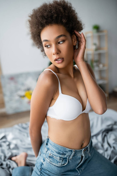sensual african american woman in white bra and jeans fixing curly hair while posing at home - Foto, Imagen