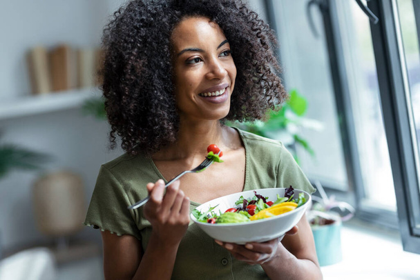 Shot of beautiful woman eating healthy salad while sitting on couch at home. - 写真・画像