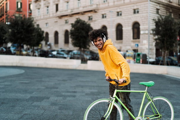 Genç Afrikalı genç adam, kulaklıkla müzik dinlerken şehirde bisikletle eğleniyor. Gençlik kuşağı yaşam tarzı ve teknoloji kavramı. - Fotoğraf, Görsel