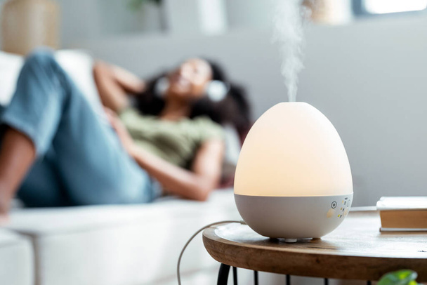 Shot of essential oil aroma diffuser humidifier diffusing water articles in the air while woman listening music lying on coach. - Fotó, kép