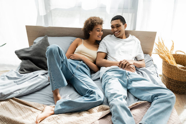 joven africano americano pareja en jeans sonriendo mientras acostado en cama - Foto, Imagen