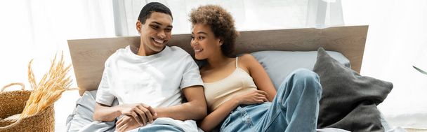 alegre afroamericana mujer acostada en la cama en casa y sonriendo a joven novio, pancarta - Foto, Imagen