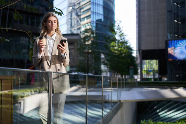 Корпоративна жінка в бежевому костюмі, п'є ранкову каву, дивиться на екран свого смартфона, використовує програму, перевіряє повідомлення по телефону, стоїть на вулиці поза офісом
. - Фото, зображення