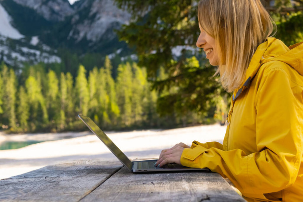 Uśmiechnięta kobieta korzystająca z laptopa z niesamowitym widokiem na góry. Wolny strzelec w żółtej kurtce i pracujący zdalnie w lesie.  - Zdjęcie, obraz