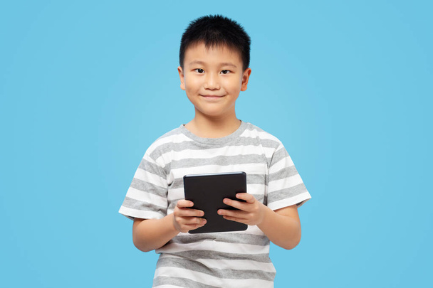 Smart kid holding tablet with smile and looking at camera, wearing t shirt on blue background - Fotó, kép