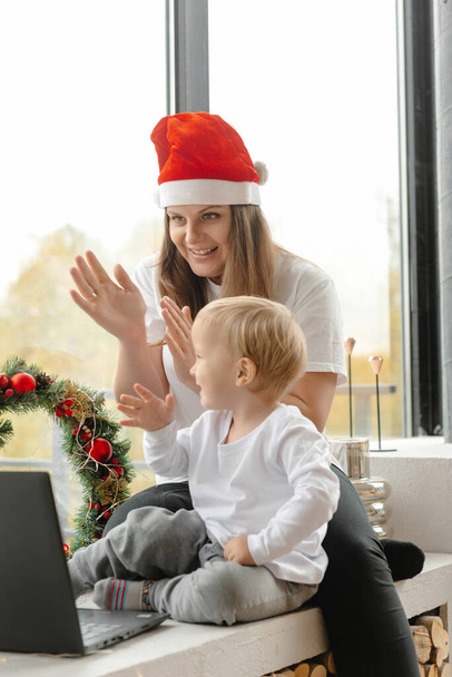 Online Χριστούγεννα. Μητέρα και γιος χαιρετούν τους συγγενείς τους μέσω μιας κάμερας laptop. Οικογένεια κάθεται μπροστά από laptop επικοινωνία μέσω video link. - Φωτογραφία, εικόνα