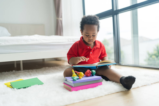 Cute little boy reading a book on a white floor in the bedroom with lots of band toys. - Photo, image