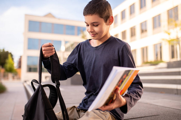 Teenage boy seated on the porch of a building lifting his backpack by the shoulder strap - Foto, Imagen