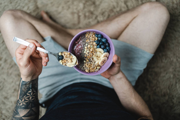Homme avec tatouage assis sur le tapis tout en mangeant un bol de granola avec smoothie, fruits et noix. Vue rapprochée du dessus. Personne méconnaissable - Photo, image