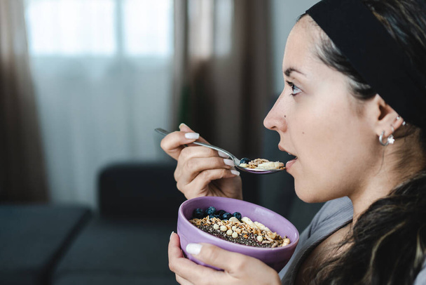 Young Latina sitting in profile eating muesli bowl for breakfast with fruits and nuts. Close up caption. Healthy lifestyle concept - Photo, Image