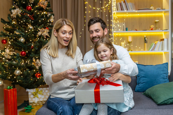 Festive portrait of a young happy family near the Christmas tree at home. Mom, dad and baby daughter are sitting on the couch and exchanging gifts, joyfully giving each other boxes with surprises. - Foto, afbeelding