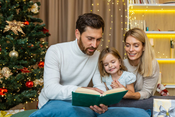 A young happy family spends time together on Christmas Eve. Mom and dad are reading a book to their daughter, sitting on the sofa near the Christmas tree. - Valokuva, kuva