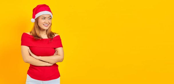 笑顔若いアジアの女性の肖像黄色の背景に手を横断クリスマス帽子を身に着けている - 写真・画像