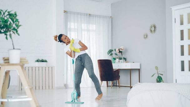 Happy African American woman is washing floor with plastic mop, singing and dancing listening to music through headphones. People and modern technology concept. - 写真・画像