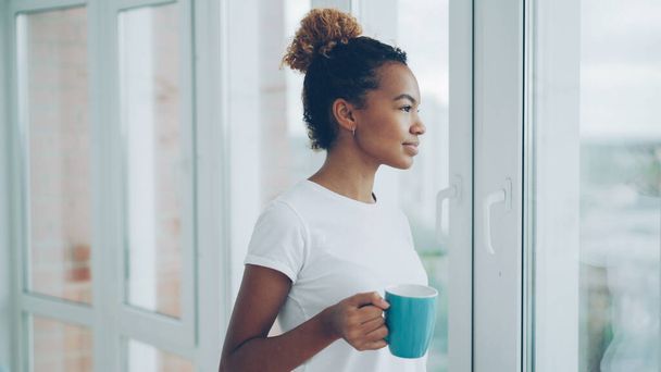 Smiling young African American woman is drinking tea standing near the window and looking outside enjoying leisure time at home. Apartments, drinks and people concept. - Фото, изображение