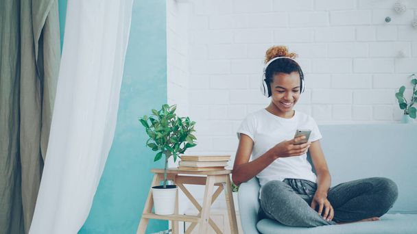 Good-looking African American girl is listening to music through wireless headphones and using smartphone sitting on sofa at home and laughing. People and technology concept. - Фото, зображення