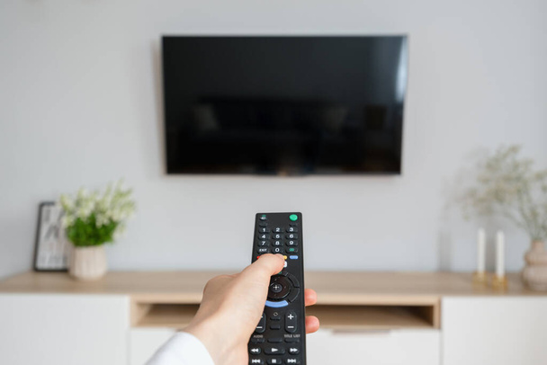 female hand hold tv remote control and press button to turn on television, sitting on couch in living room at cozy apartment, entertainment and relax at home - Photo, Image