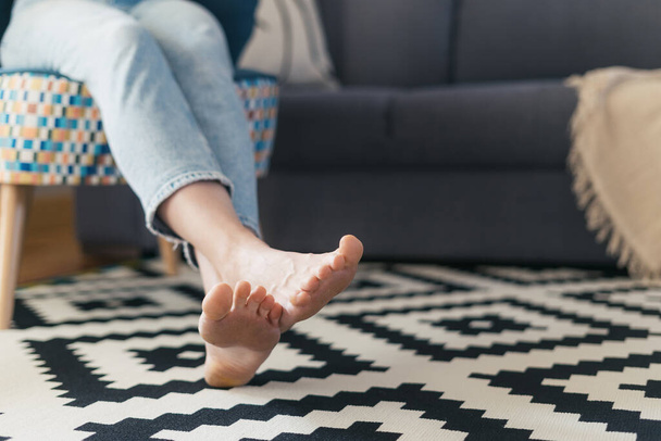 cropped shot of female with barefoot legs on floor carpet relaxing on comfort armchair near couch in living room at cozy apartment - Zdjęcie, obraz