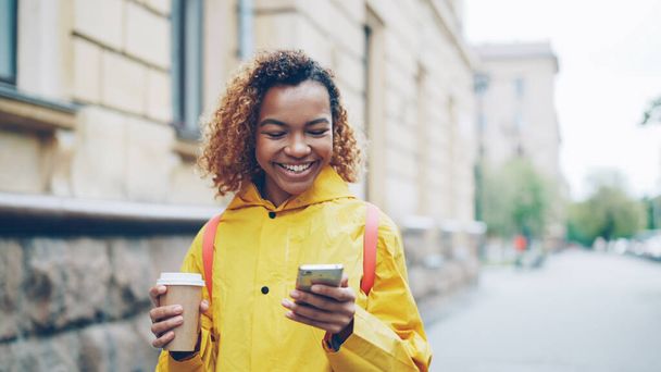 Happy African American woman is chatting online with friends using smart phone, smiling and holding take out coffee. Beautiful city with green trees and old buildings is visible. - Foto, Imagem