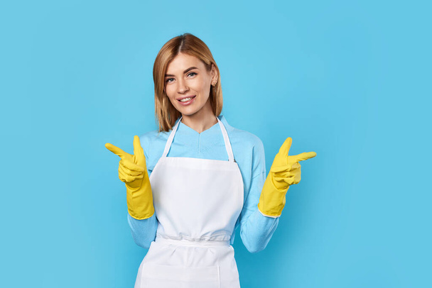 beautiful caucasian blonde woman in yellow rubber gloves pointing fingers at copy space isolated on blue background. cleaning - Zdjęcie, obraz