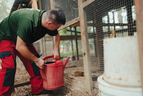 Caretaker with down syndrome in the zoo giving water in animal enclosure. Concept of integration people with disabilities into society. - 写真・画像
