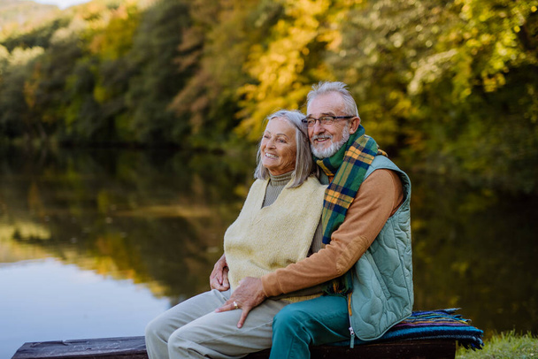 Senior couple in love sitting together on a bench near lake, during autumn day. - Zdjęcie, obraz