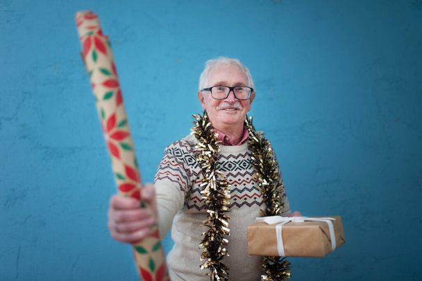 Portrait of happy senior man holding a Christmas gift and wrapping paper studio shooting. - Photo, Image