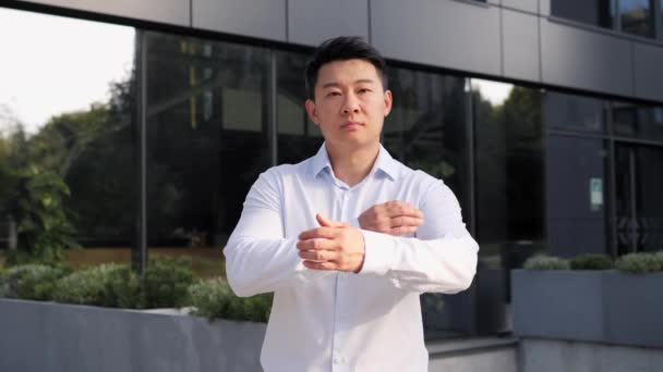 Portrait of young adult serious asian businessman looking at the camera while folding hands outdoor. Successful male business man standing on the background of modern grey glass office center - Footage, Video