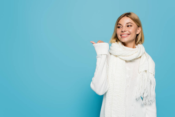 cheerful blonde woman in white sweater and scarf pointing away with thumb isolated on blue  - Foto, imagen