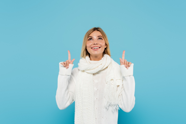 fröhliche blonde Frau in weißem Pullover und Schal mit nach oben zeigenden Fingern auf blauem Grund  - Foto, Bild