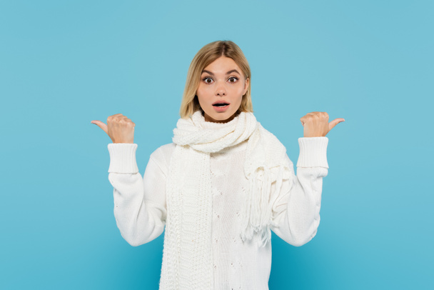 Verwirrte blonde Frau in weißem Pullover und Schal, die mit Daumen auf blau zeigt  - Foto, Bild