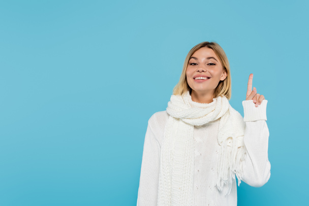 cheerful blonde woman in white sweater and scarf pointing up with finger isolated on blue  - Foto, Imagem