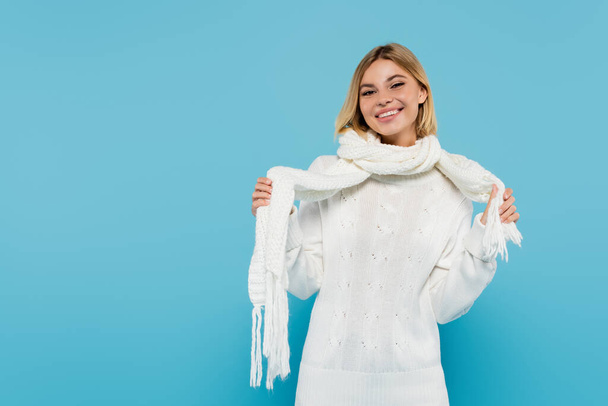 happy blonde woman in white sweater adjusting winter scarf isolated on blue  - Photo, Image