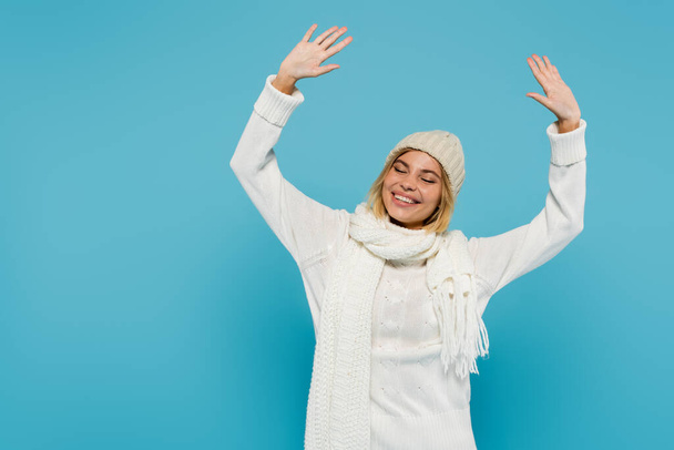 femme blonde positive en pull blanc et chapeau d'hiver levant les mains isolé sur bleu - Photo, image