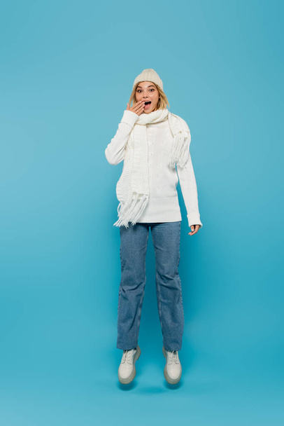 volle Länge der aufgeregten jungen Frau im winterlichen Outfit, die den Mund bedeckt, während sie auf blau schwebt  - Foto, Bild