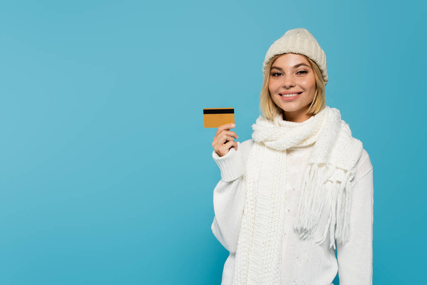 mujer rubia feliz en suéter blanco y sombrero de invierno con tarjeta de crédito aislada en azul - Foto, imagen