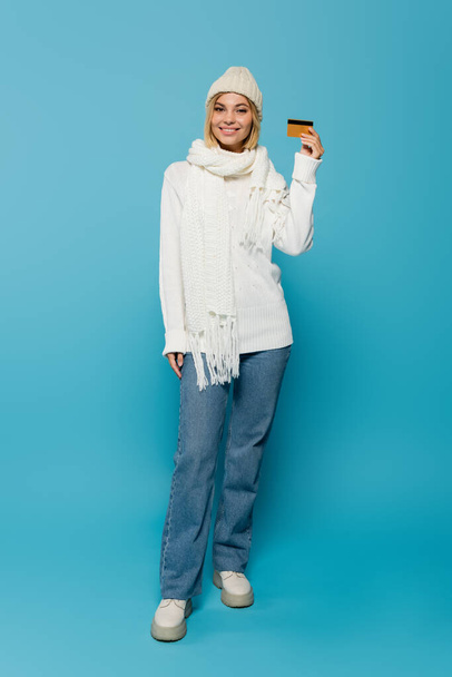 full length of happy blonde woman in white sweater and winter hat holding credit card on blue - Foto, imagen