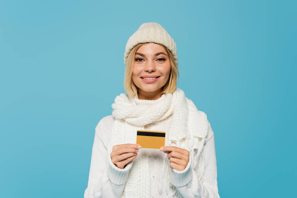 mujer alegre en suéter blanco y sombrero de invierno que sostiene la tarjeta de crédito aislada en azul - Foto, imagen