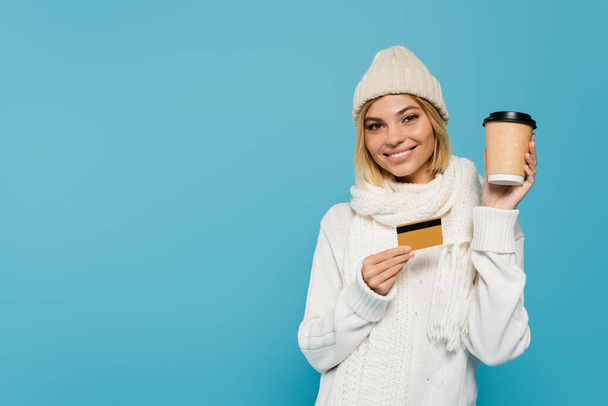 счастливая блондинка в белом зимнем наряде держа кредитку и бумажный стаканчик изолированы на синий - Фото, изображение